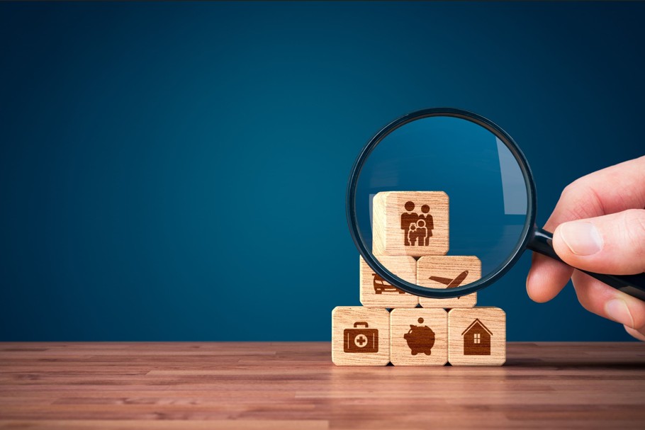 Une Main Tient Une Loupe Au Dessus De Blocs De Bois Empilés Sur Une Table, Chaque Bloc Affichant Une Icône Représentant Les Finances, La Santé, La Famille, L'épargne Et La Maison.