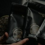 Gros Plan Sur Des Mains Tenant Des Paquets Noirs étiquetés "chocolate Mint", "gorilla" Et "watermelon", Mettant En évidence Les Variétés D'intérieur Et D'extérieur Disponibles Pour L'achat De Cbd En Ligne.