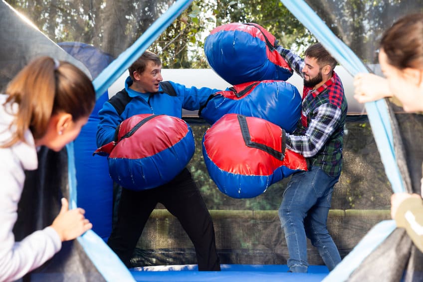 Deux Hommes Portant Des Gants De Boxe Gonflables Surdimensionnés S'amusent à S'affronter à L'intérieur D'un Espace Clos Tandis Que Deux Personnes Les Observent De L'extérieur.