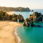 Aerial view of Golden Bay, New Zealand, featuring golden sand, turquoise water, dramatic rock arches, and lush green hills under a warm glow. A person walks on the beach.