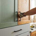 A hand installs a matte black handle on a wooden kitchen cabinet, next to a freshly painted sage green cabinet and grey drawer.