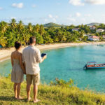 A couple stands on a grassy viewpoint overlooking a turquoise Martinique bay with a white sand beach, palm trees, a fishing boat, and colorful houses.