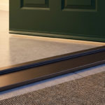 Low-angle view of a dark green front door with a new, brushed dark metal threshold and subtle weatherstrip. Warm interior light illuminates the concrete floor and doormat.