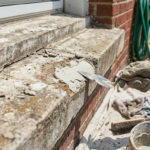 Close-up of gloved hands applying repair mortar to a damaged concrete window sill of a brick house, in bright daylight.