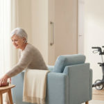 Smiling senior woman in a sunlit living room reaching for a book. A modern rollator and grab bar are subtly integrated.