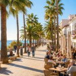 A vibrant, sun-drenched Spanish coastal promenade lined with palm trees and cafes, with people strolling and dining, overlooking the sparkling blue sea.