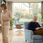 Seniors dans un salon lumineux, la femme gère la maison connectée via une tablette, l'homme lit. Confort moderne et autonomie.