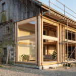 A rustic barn undergoing renovation, with weathered wood contrasting new timber frames, large windows, and modern interior glimpses, scaffolding visible.
