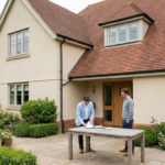 Deux hommes discutant un accord devant une grande maison moderne avec jardin bien entretenu, documents sur une table.