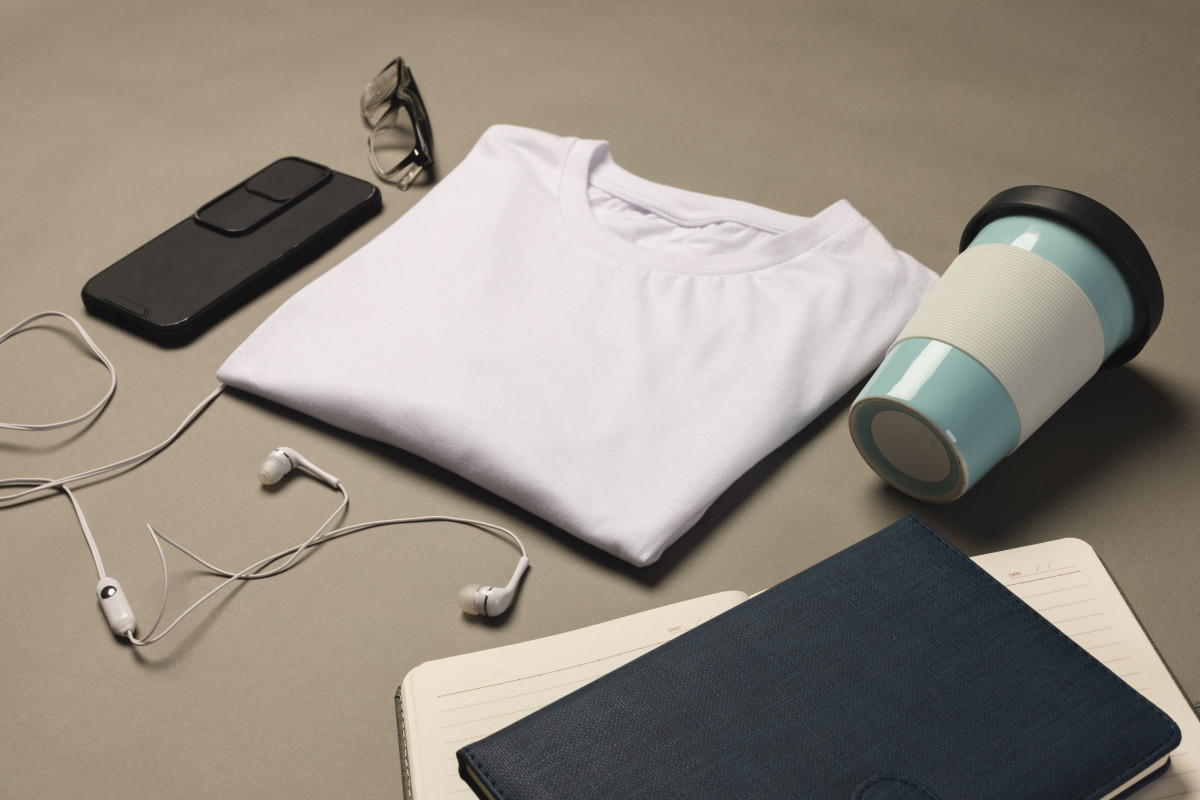 Vue à Plat D'un T Shirt Blanc Plié, D'un Gobelet Réutilisable, De Lunettes, D'un Smartphone, D'écouteurs, D'un Carnet De Notes Et D'un Journal Sur Un Fond Neutre.