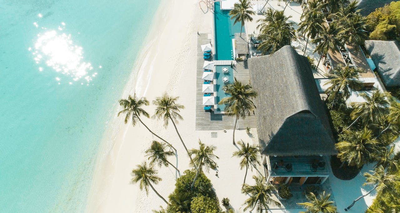 Vue Aérienne D'une Station Balnéaire Tropicale Avec Des Palmiers, Une Piscine, Des Parasols Et L'eau Claire Et Turquoise De L'océan Le Long D'un Rivage De Sable Blanc.