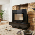 A worker inspects a partially reconstructed brick chimney breast with a flashlight, installing a sleek black fireplace insert in a modern living room. Renovation tools are in the foreground.