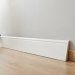 Mains gantées posant une surplinthe blanche profilée sur une ancienne plinthe en bois, avec outils de rénovation.