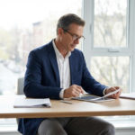 Mature man in business attire at a desk with tablet and papers, bathed in daylight from a window overlooking a cityscape.