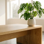 Plante Monstera verte en pot blanc sur une longue table en bois clair dans une pièce moderne et lumineuse avec fenêtres.